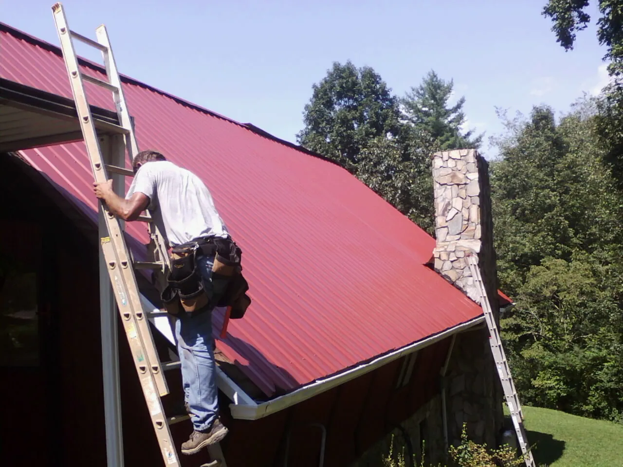 Roofer on a ladder beside a red metal roof and stone chimney — Soffit & Fascia Repair by Roofing Brothers in Miamisburg